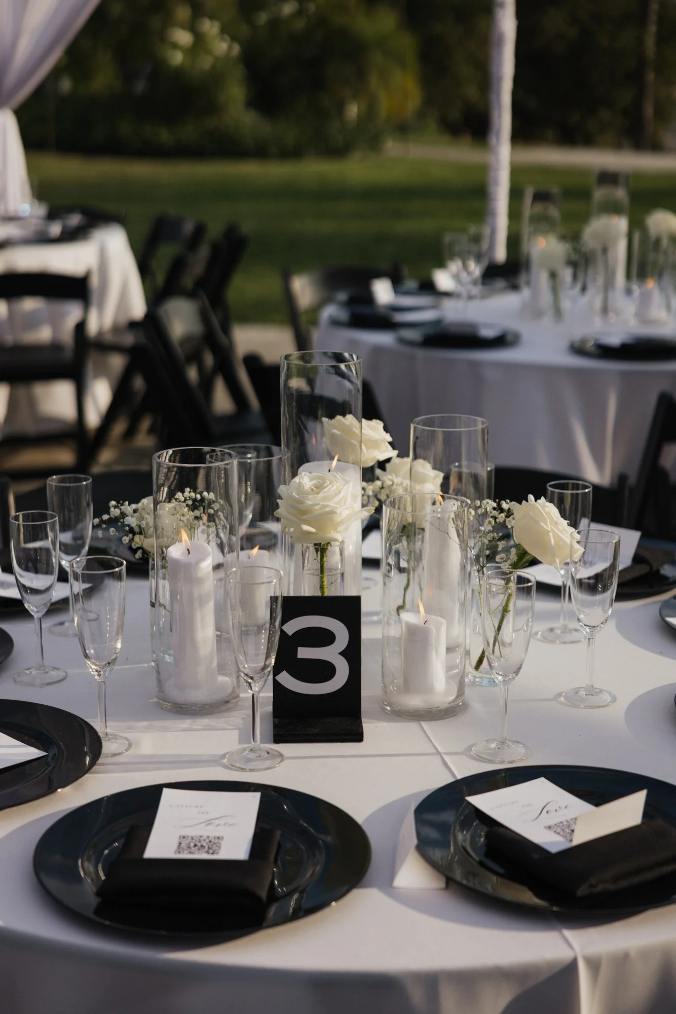 Elegant outdoor wedding reception table setup with white roses in glass vases, candles, and black plates with black napkins, numbered table card, and QR codes on napkins, with trees and grass in the background at the Grove Redlands in California