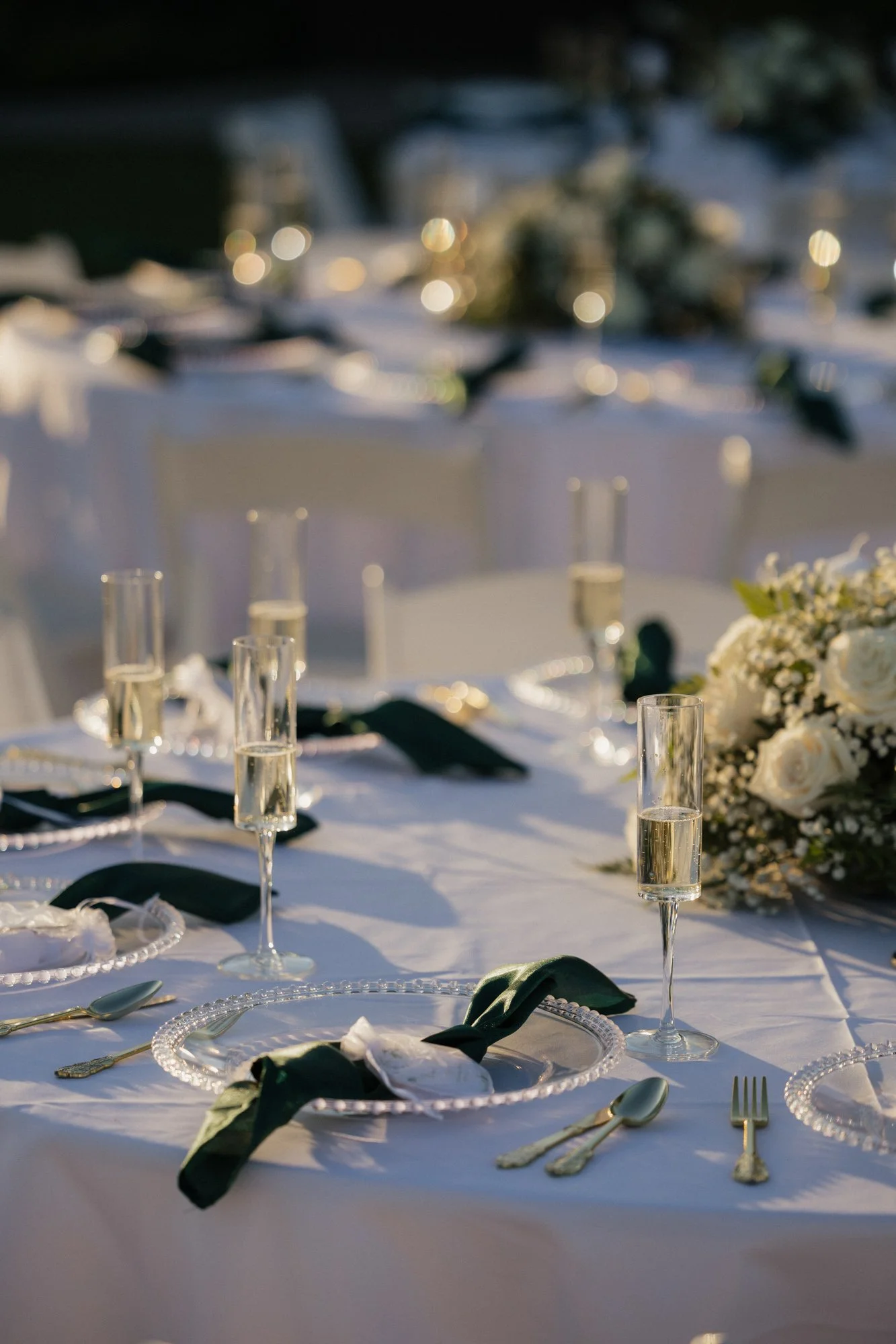 Elegant outdoor wedding reception table set with white tablecloth, champagne glasses, floral centerpieces, white plates, gold utensils, and dark green napkins, illuminated by warm lighting in Redlands California