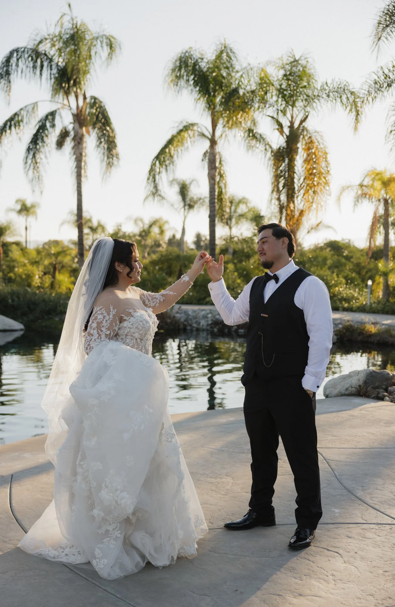 A bride and groom outdoors near a pond, with palm trees in the background, holding hands and facing each other on their wedding day at the Grove redlands in Southern California 