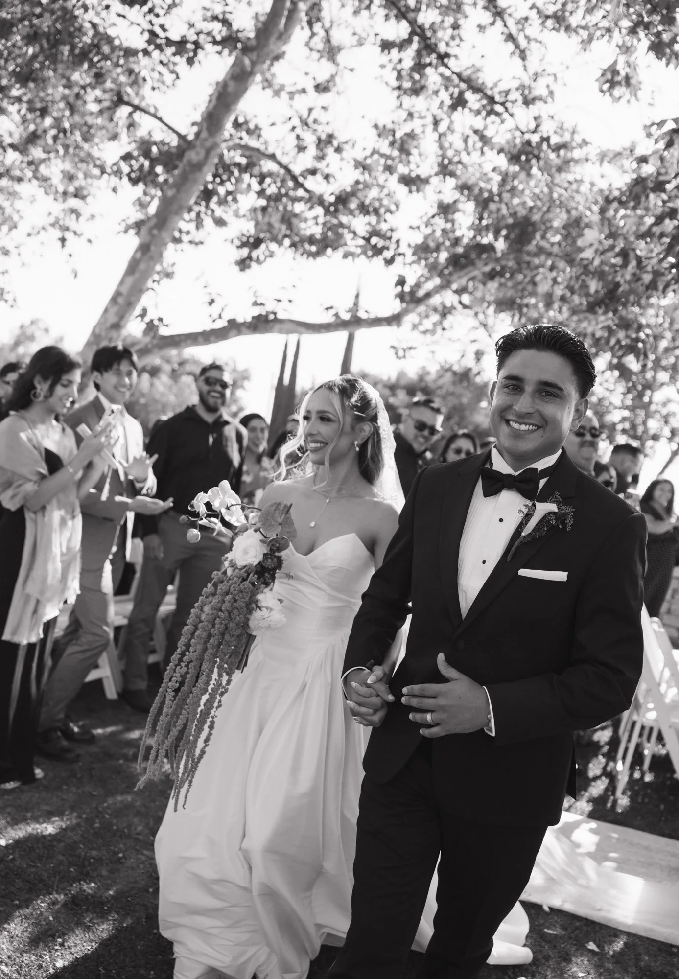 Candid photo of a wedding scene showing a bride and groom holding hands and smiling after ceremony, surrounded by friends and family outdoors under trees in Diamond Bar California