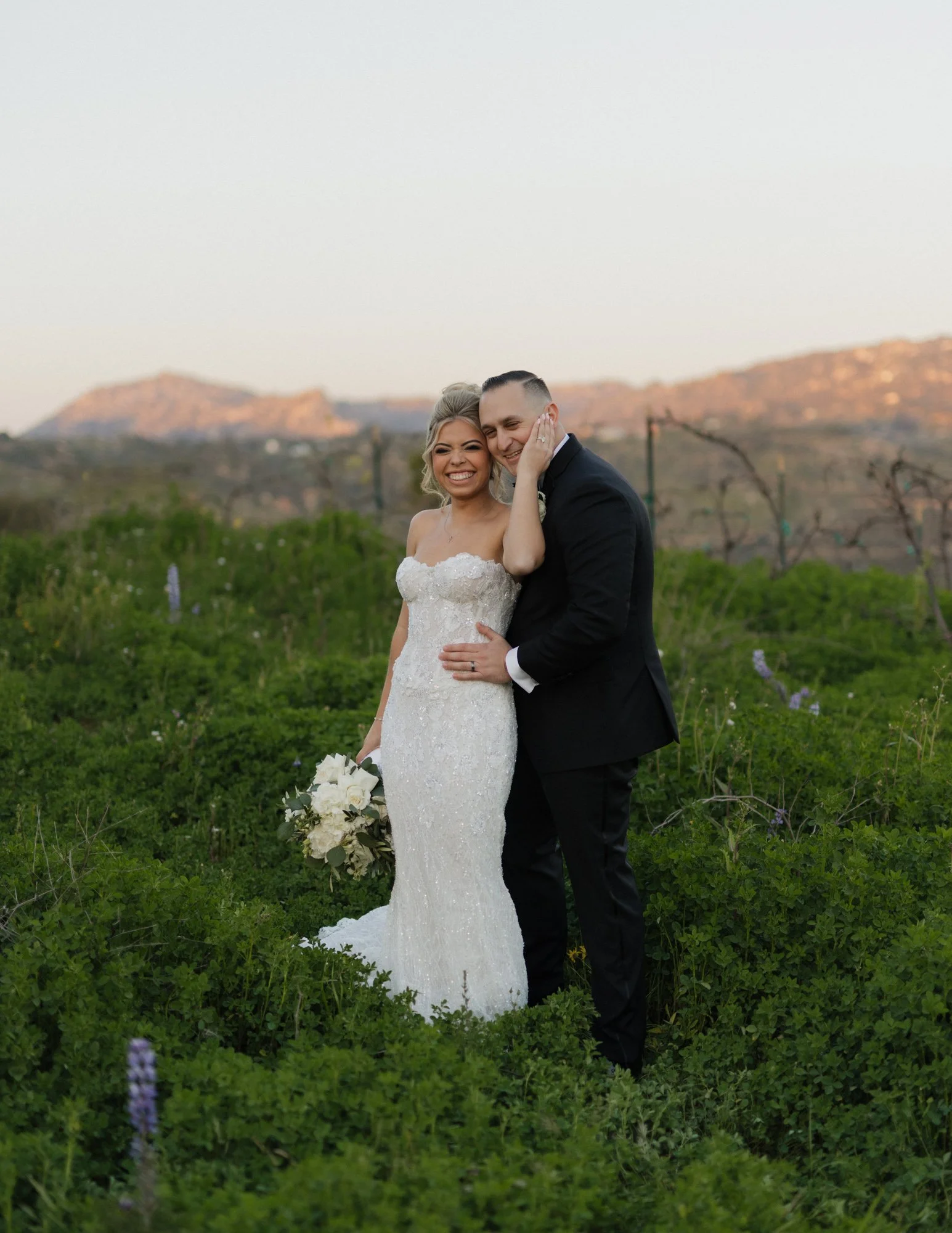 A happy bride and groom embracing outdoors, with a scenic background of mountains and green fields, during sunset or golden hour at Tivoli wedding venue in Fallbrook California.