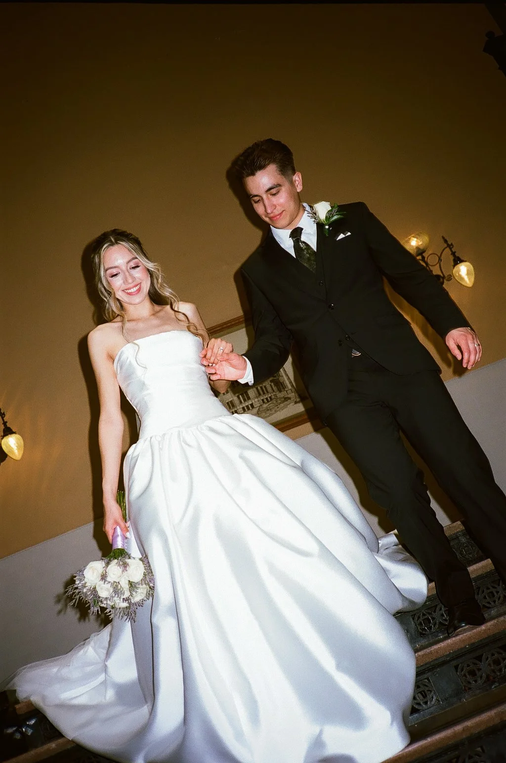 A bride in a white wedding gown holding a bouquet and a groom in a black suit walking down a staircase, smiling and looking down in at Old Orange County Courthouse.