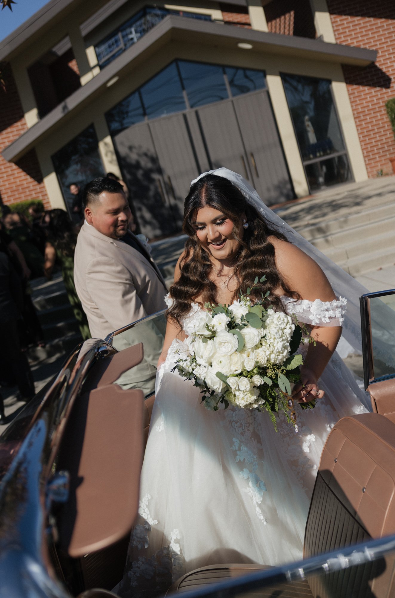 A candid photographer takes a candid photo of groom helping bride get into a vintage car on their wedding day in Chino Hills California.