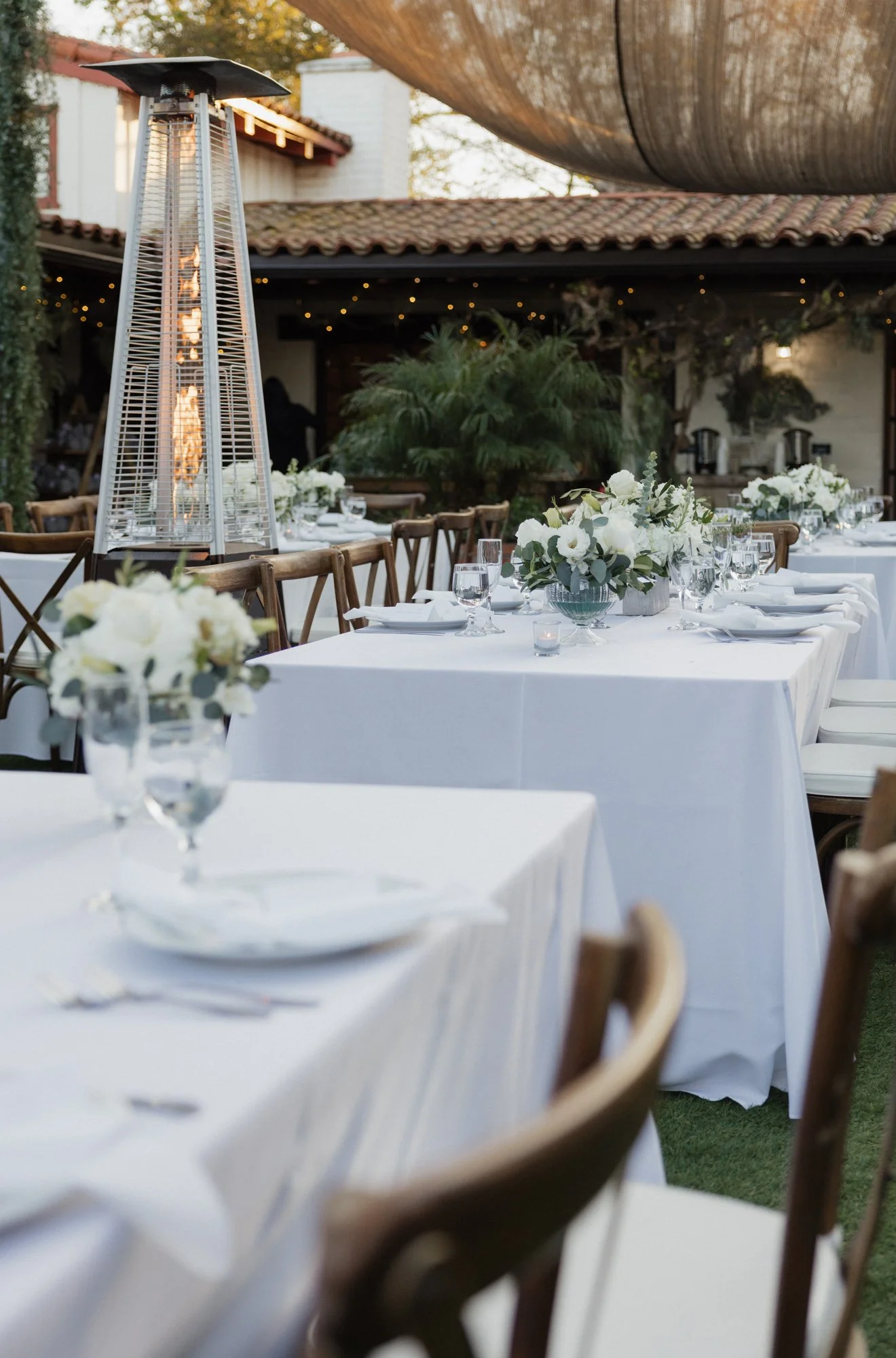 Outdoor dining setup with white tablecloths, floral centerpieces, glassware, and wooden chairs, with a patio heater and a shade structure overhead at Tivoli wedding venue in Fallbrook California.