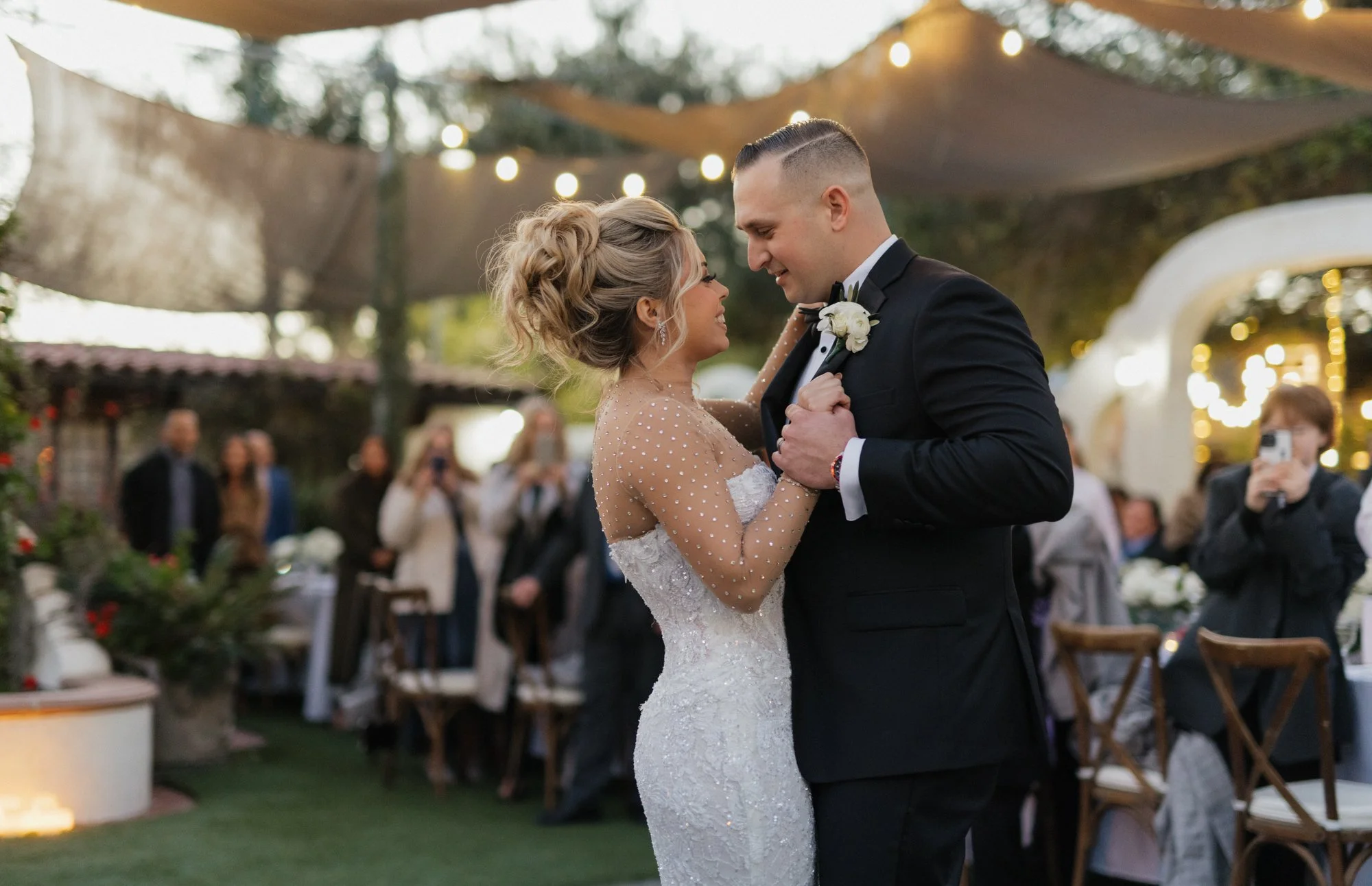 A bride and groom dance closely at their wedding reception outdoors, surrounded by guests and string lights in a decorated garden setting at Tivoli wedding venue in Fallbrook California.
