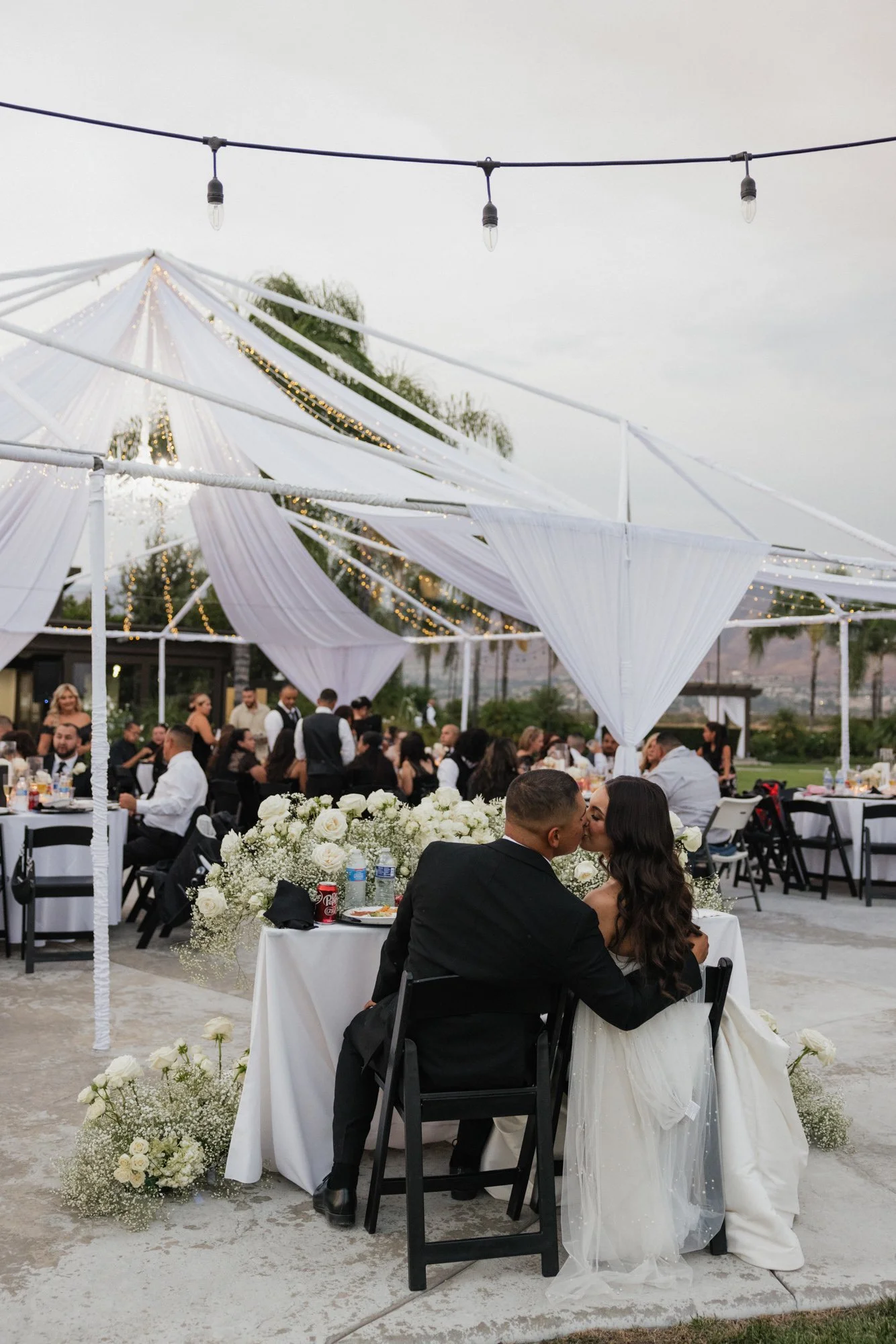 A candid moment at wedding reception outdoors with the bride and groom sitting closely and kissing at a table decorated with white flowers. Guests are seated at round tables in the background at The Grove Redlands