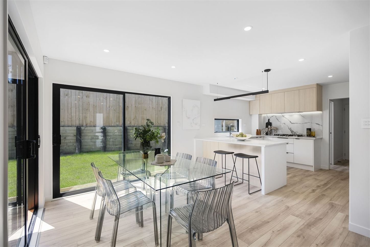 Modern kitchen and dining area with glass table, transparent chairs, wooden flooring, large windows overlooking a backyard, and a kitchen island with stools.