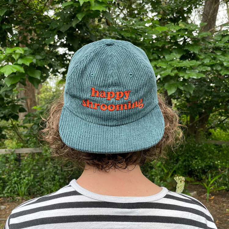 Clare wears a pine green corduroy cap with the words 'Happy Shrooming' embroidered in burnt orange.