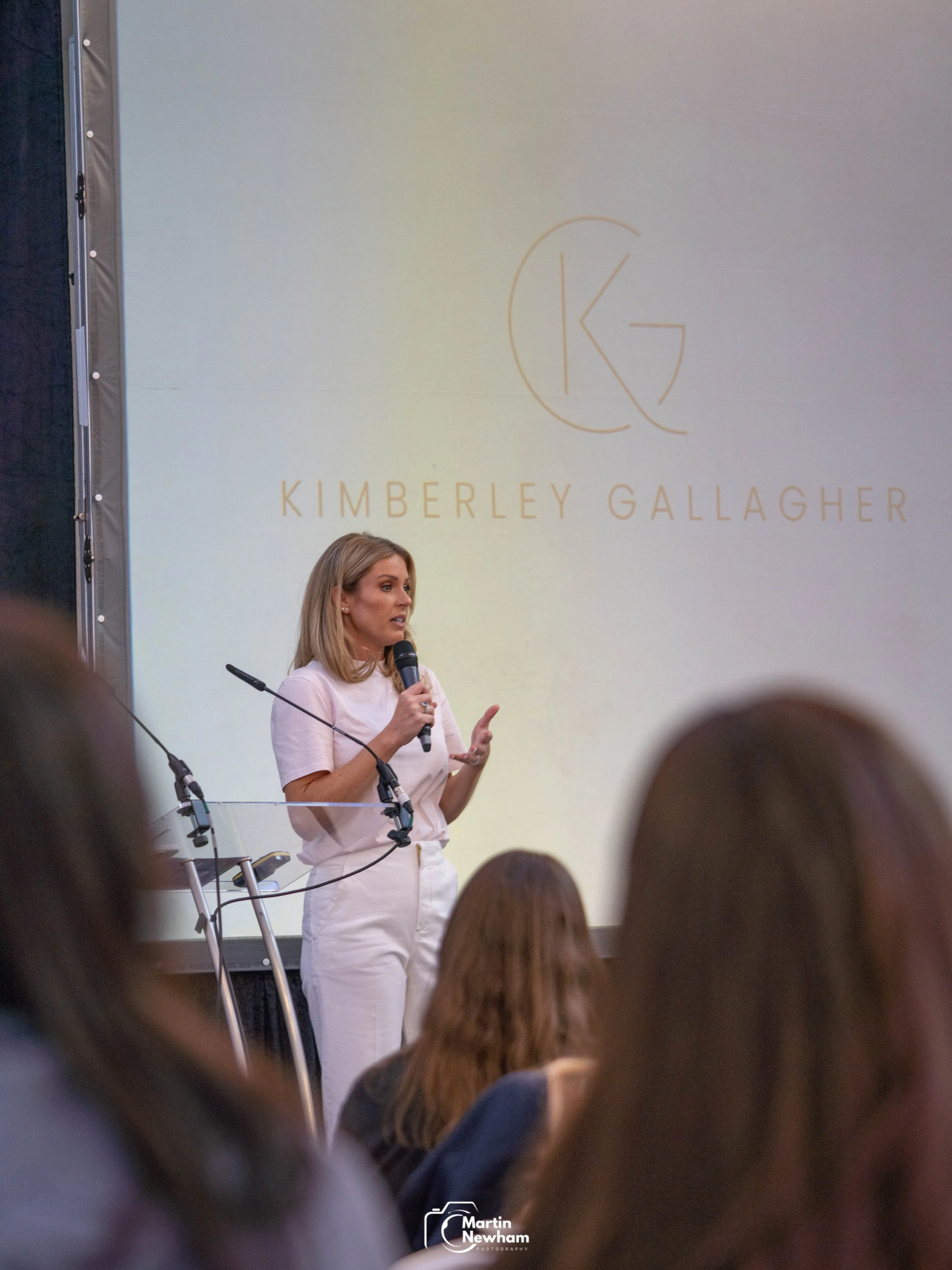 A woman with blonde hair, wearing a white outfit, is standing on stage with a microphone, reading from a piece of paper. Behind her, there is a large decorative blue backdrop with white patterns and the word 'LOVE.' A screen on the left shows a close-up of a person holding a booklet and wearing a white blazer. Audience members are visible in the foreground.