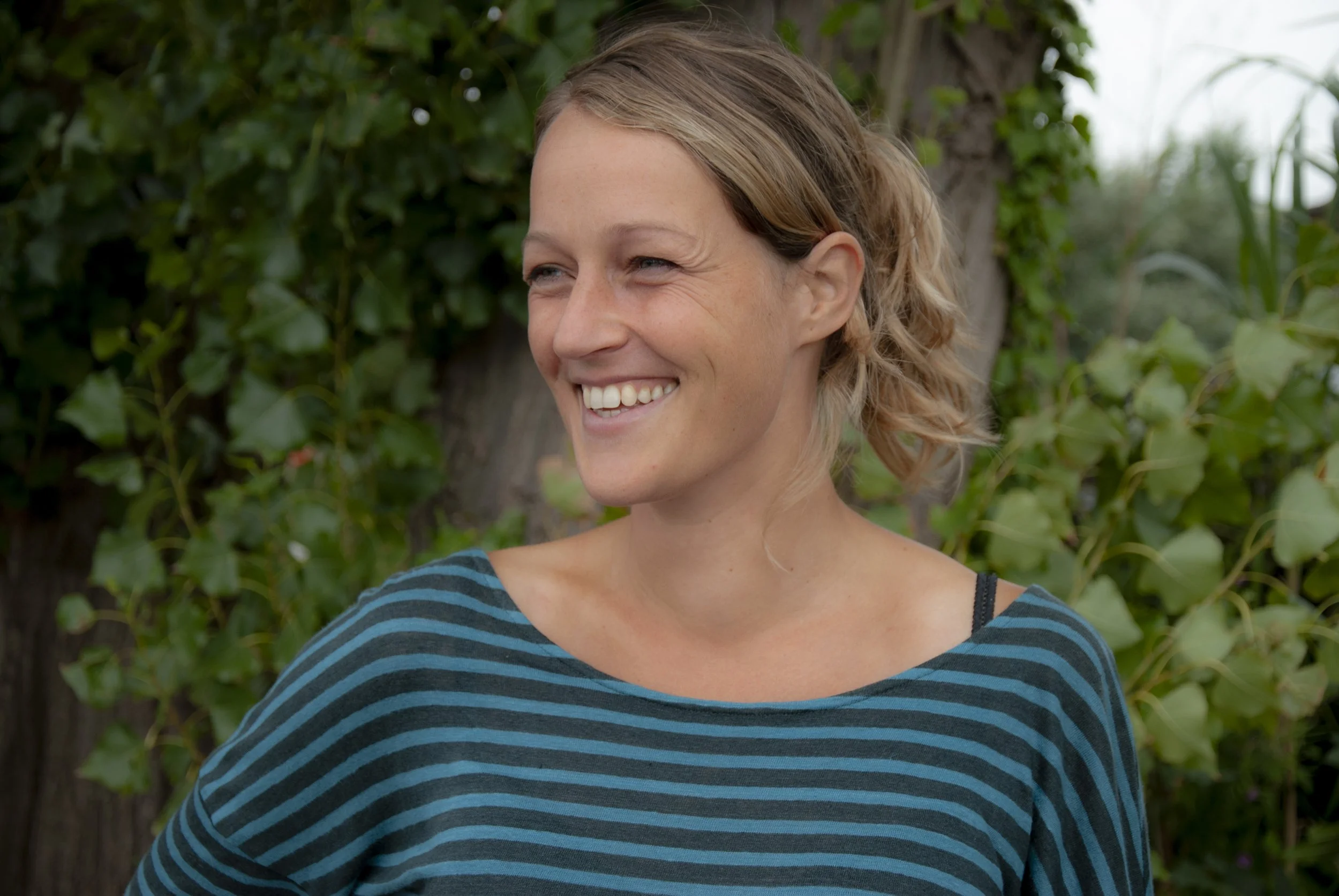 Een vrouw met blond haar draagt een beige blazer en een zwarte top, en heeft een kleurrijke ketting en oorbellen, terwijl ze buiten staat met een bos van bomen op de achtergrond.