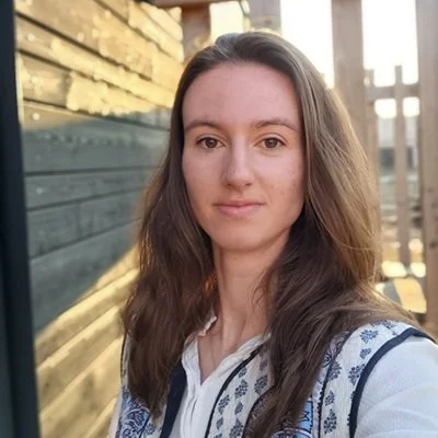 Portret van een jonge vrouw met bruin, golvend haar, staande buiten met een houten muur en een terras in de achtergrond.