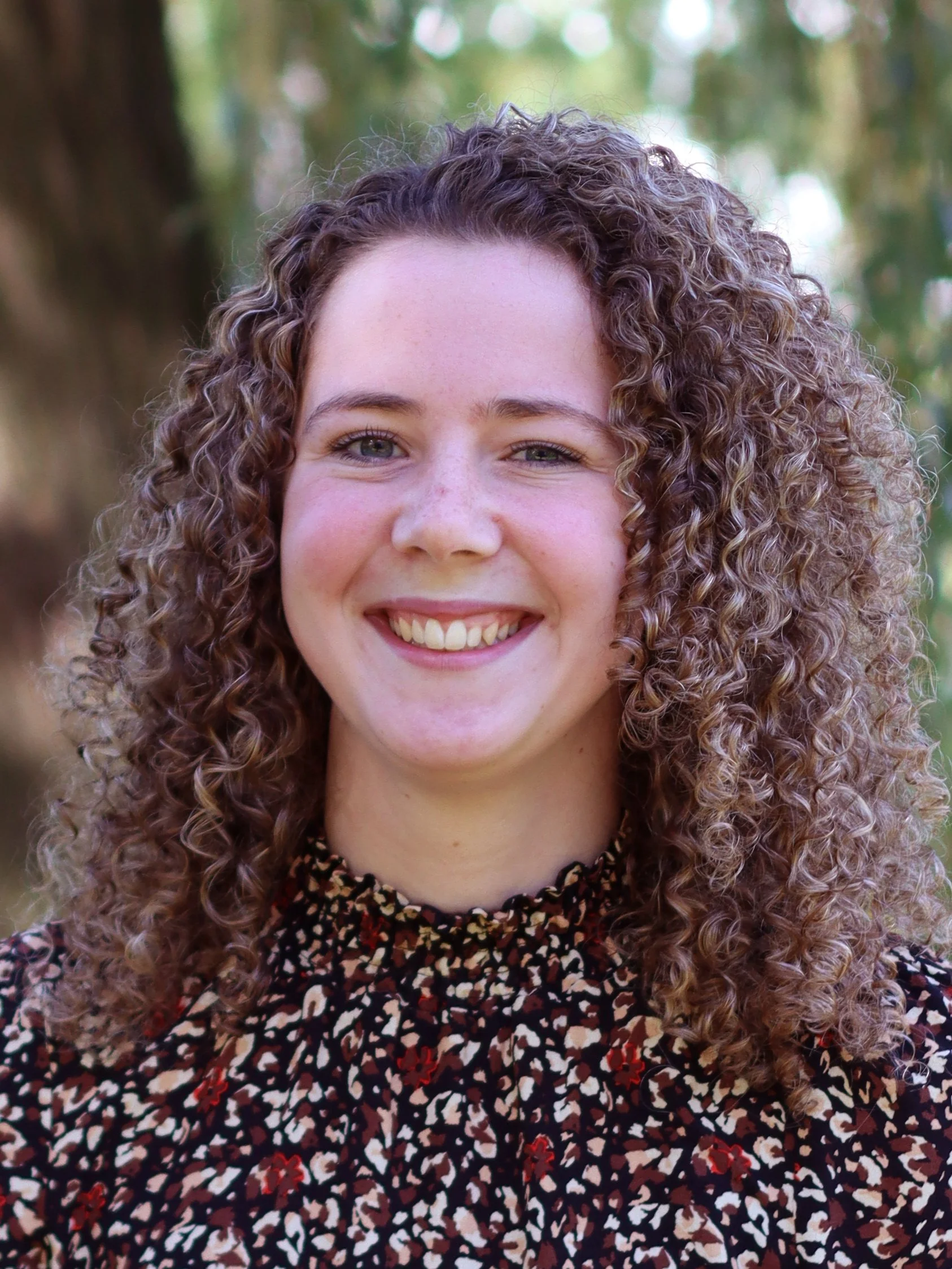 Portret van een lachende vrouw met gekruld, bruin haar, buiten met groene achtergrond.