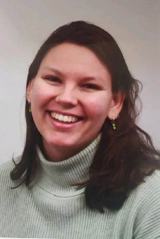 Close-up van een lachende vrouw met bruin haar, draagt een lichtgrijze trui en oorbellen, tegen een witte achtergrond.