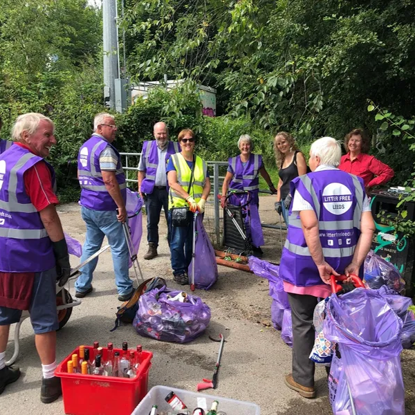 Group of volunteers wearing purple and yellow vests picking up litter along a sidewalk, holding trash bags and tools, with bags of collected trash on the ground.