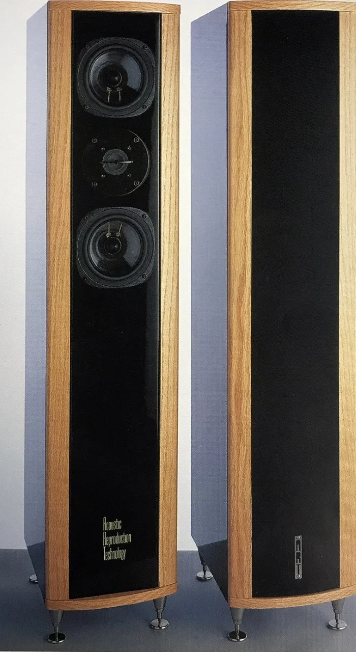 Two tall wooden-frame speakers standing on small metal legs, with the left speaker displaying two large drivers and a mid-range driver, and the right speaker having a black grill covering the drivers. The left speaker has text reading 'Acoustic Reproduction Technology' near the bottom.