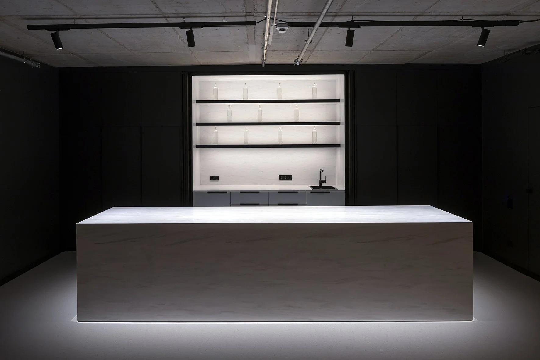 Projekt: Event Lab in Berlin. Inhalt: Moderner, minimalistischer Küchenraum mit großen weißen Marmorinsel in der Mitte, dunklen Wänden und offener Backstein-Decke, beleuchtet von schwarzen Deckenleuchten.