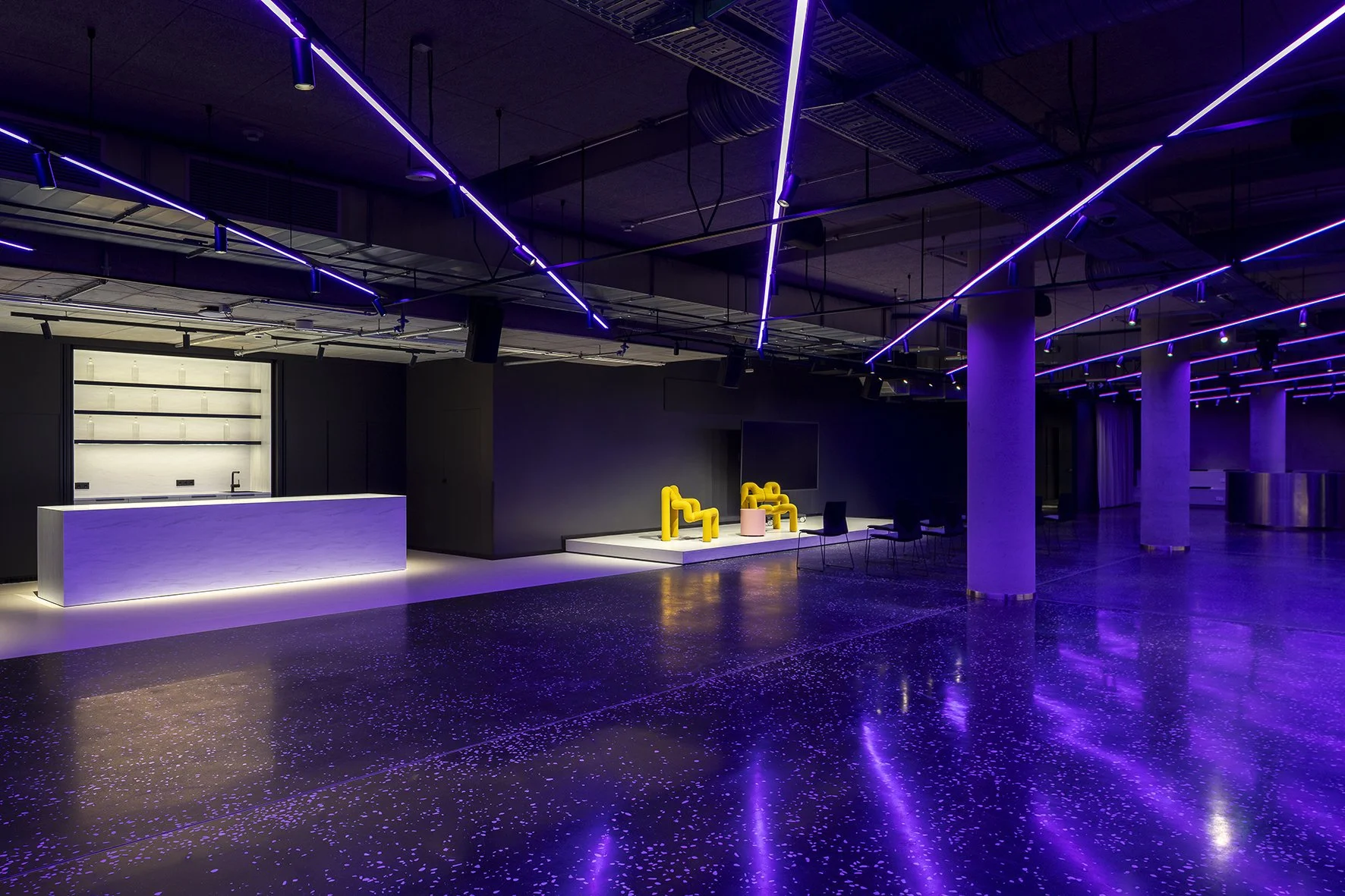 Projekt: Event Lab in Berlin. Inhalt: Ein dunkler Veranstaltungsraum mit blauer LED-Beleuchtung, eine Bar mit weißer Oberfläche, gelbe Sitzmöbel und ein großer Bildschirm, die auf einer kleinen erhöhten Plattform stehen.