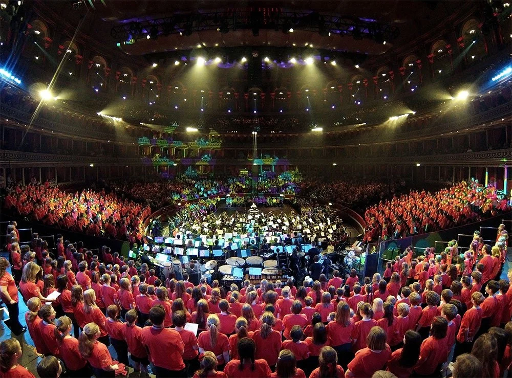 Royal Albert Hall, London: Large indoor concert hall filled with choir members wearing red robes and a symphony orchestra performing on stage, with colourful lighting and audience in seats.