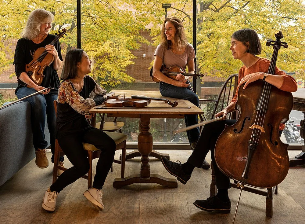 Park Theatre: Four women in a room with large windows and autumn trees outside, playing string instruments, including violins and a cello, and smiling at each other. Musician photography in London.