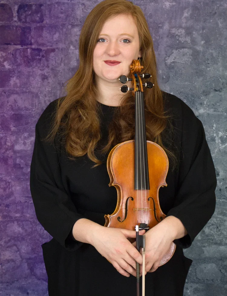 Georgina Hannant, Violin: A woman with long hair holding a violin and bow, standing in front of a brick background. Musician photography, London.