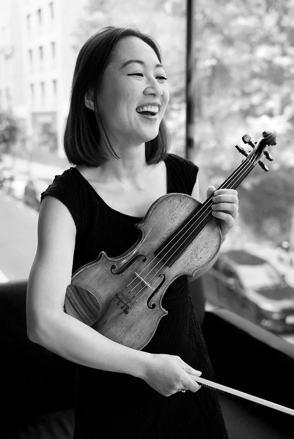 A woman holding a violin and bow, smiling, with trees and buildings visible outside in the background. Musician photography, London.