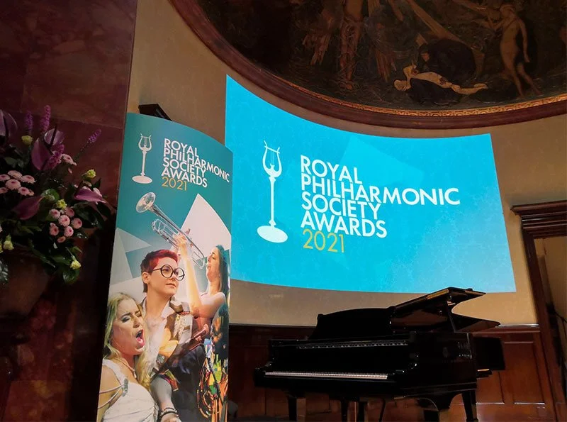 Wigmore Hall, London: Stage setup for the 2021 Royal Philharmonic Society Awards with a large screen displaying the event's logo, a seating piano, and a decorative floral arrangement.
