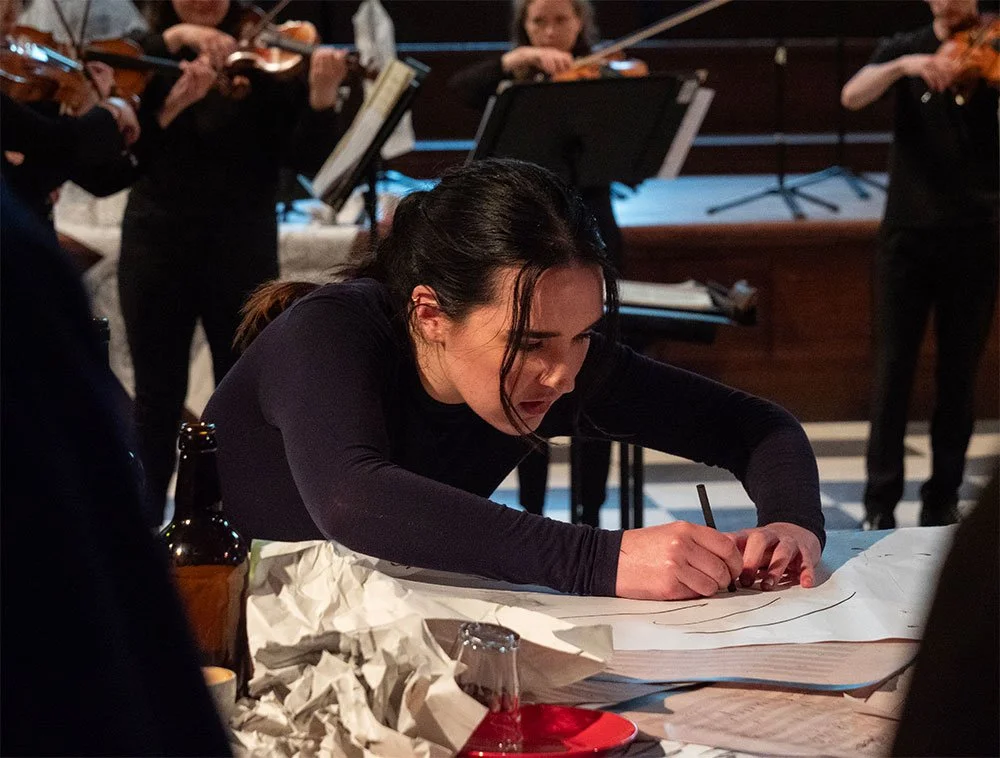 A woman sitting at a table drawing on large sheets of paper with a marker, with an orchestra playing in the background.