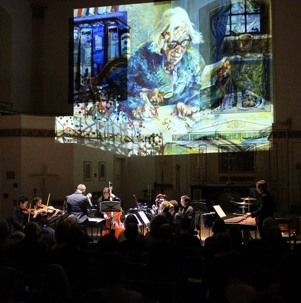 An orchestra performing in a concert hall with a large digital projection of a painting of an elderly woman reading a book projected on the wall behind them. Orchestra concert photography, London.