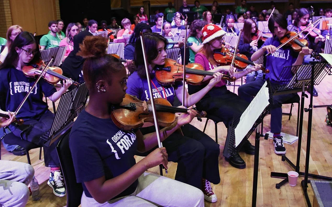 Orchestras for All: A group of young musicians playing violins during a rehearsal or concert on a stage.
