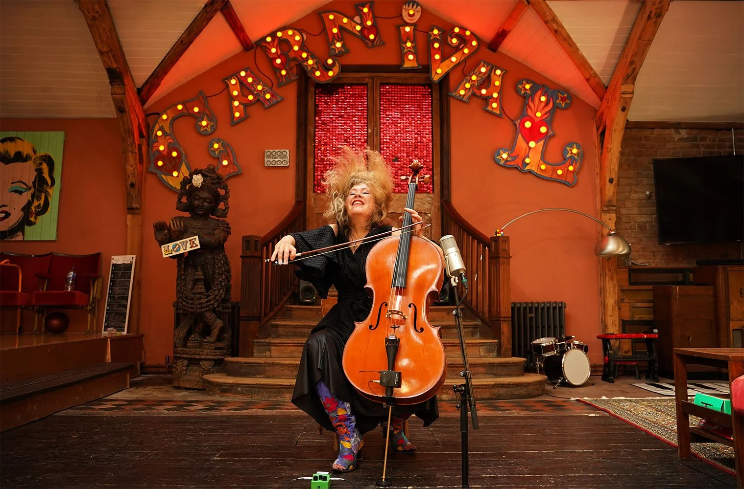 Zoe Martlew, Cellist: A woman playing a cello on stage in a colourful venue with decorative signs, artwork, and musical instruments. Musician photograph taken on-location as part of a film shoot.