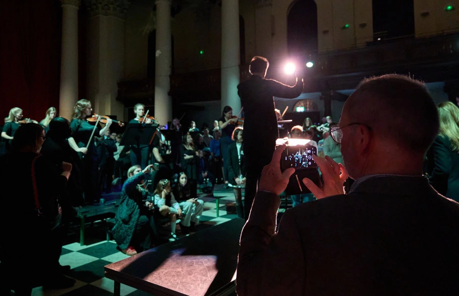 A conductor leading an orchestra during a performance with audience members watching and a person in the foreground taking a photo with a smartphone in a dimly lit concert hall.