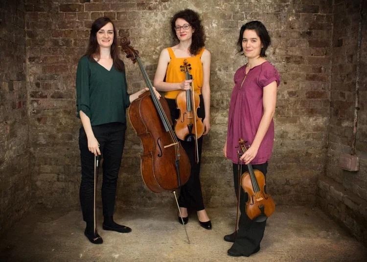 Three women holding string instruments with a brick wall background. Musician photography, London.