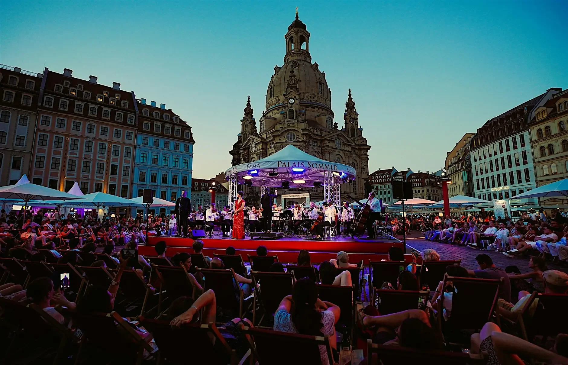 An outdoor concert in a city square at dusk, with a large crowd seated in front of a stage where a band is performing. The stage is set against historic buildings and a prominent church tower in the background. There are umbrellas and lights illuminating the scene.