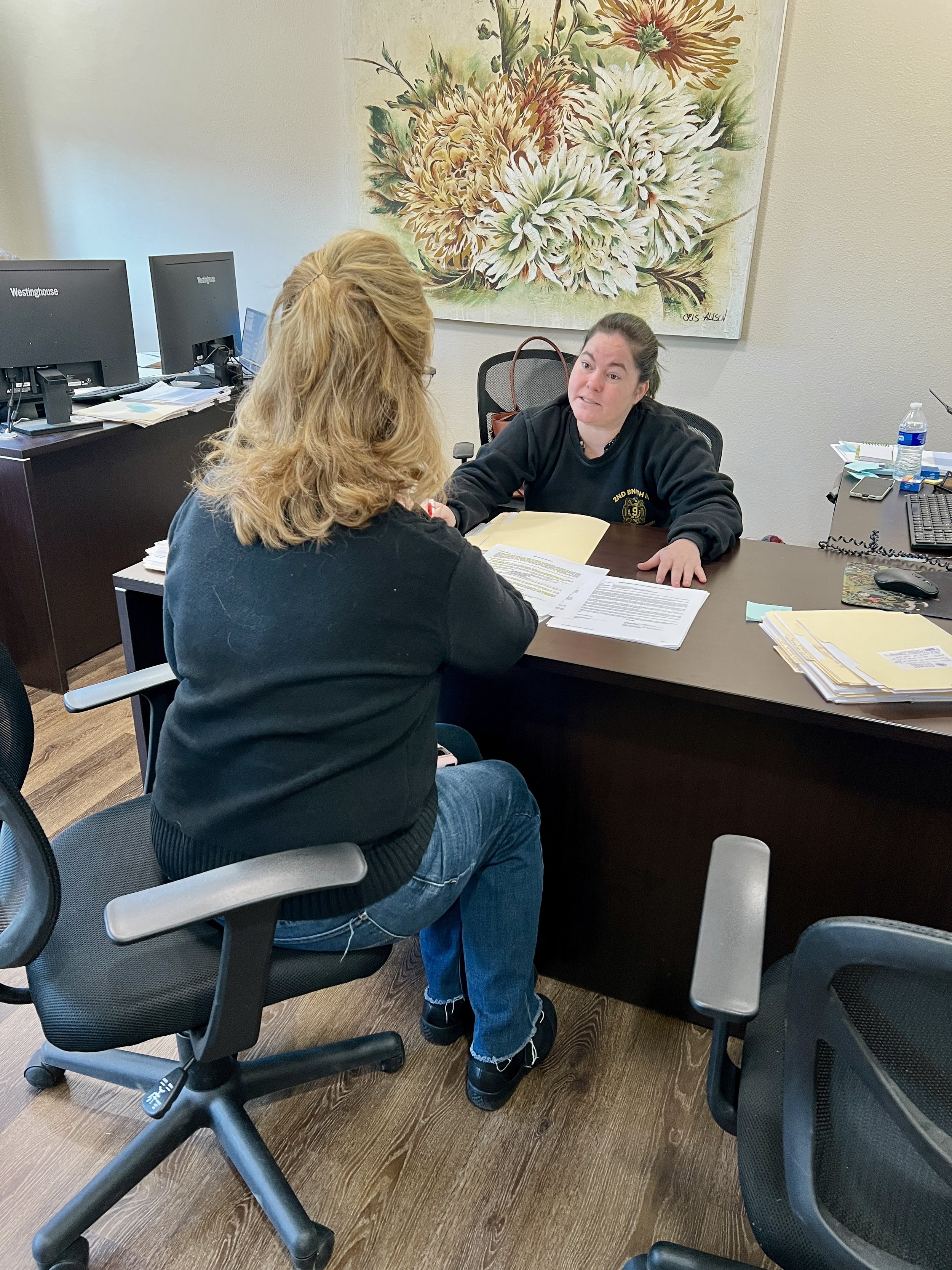 A blond woman sitting across the table from a brunette woman with paperwork and manilla file folders between them in an office setting