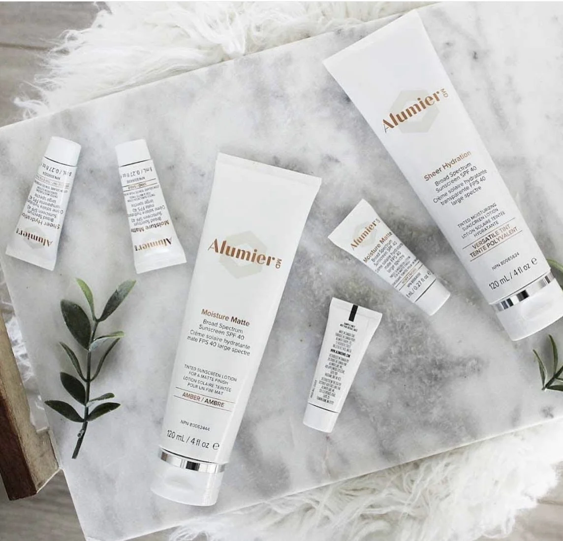 Collection of Alumier skincare products scattered on marble surface with green leafy sprig and white fluffy rug in background.