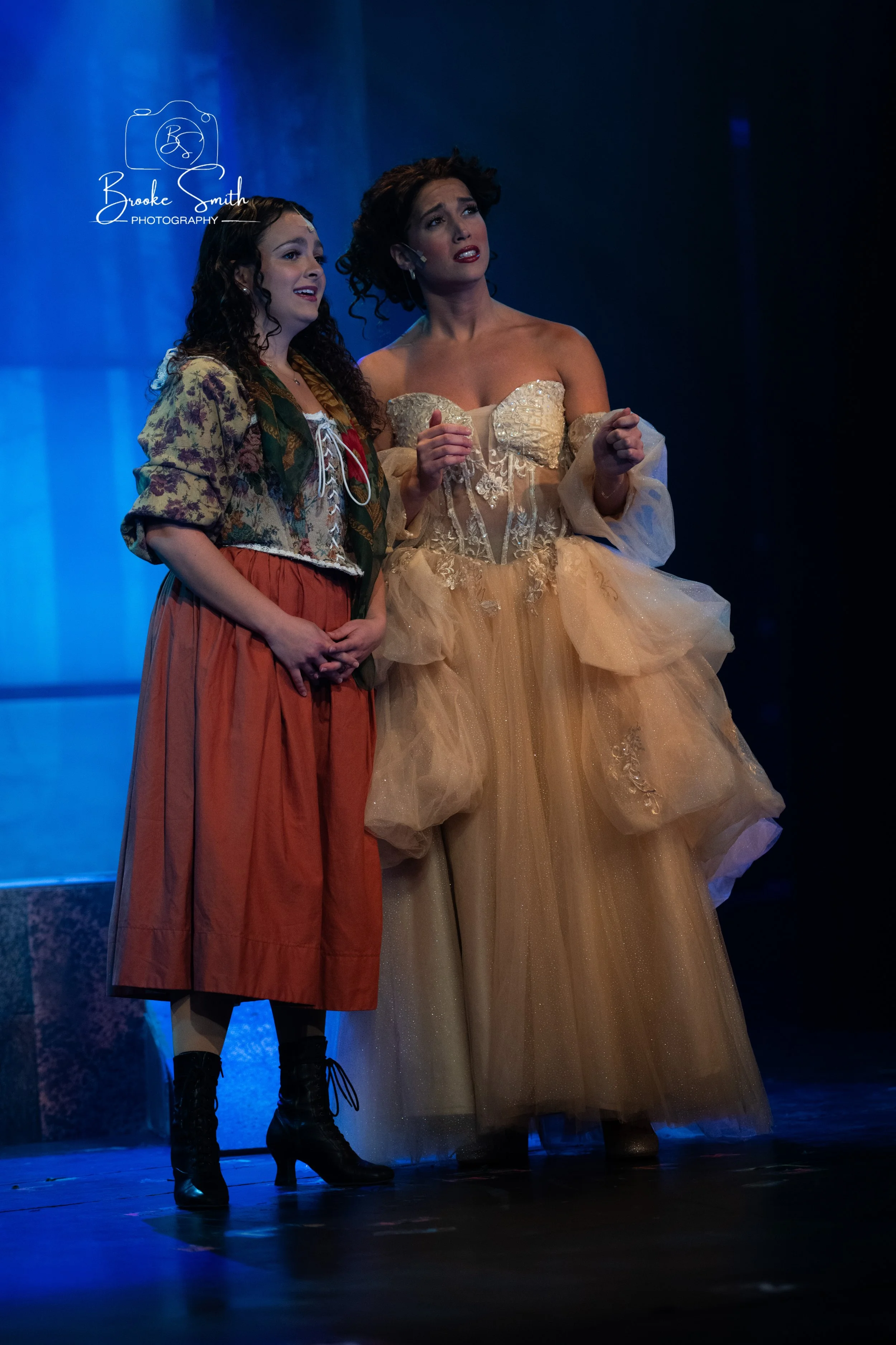 Two women stand on stage under blue lighting, one in a vintage cream ball gown with off-shoulder puffed sleeves, the other in a casual patterned jacket and rust-colored skirt, appearing to perform or act in a theatrical scene.