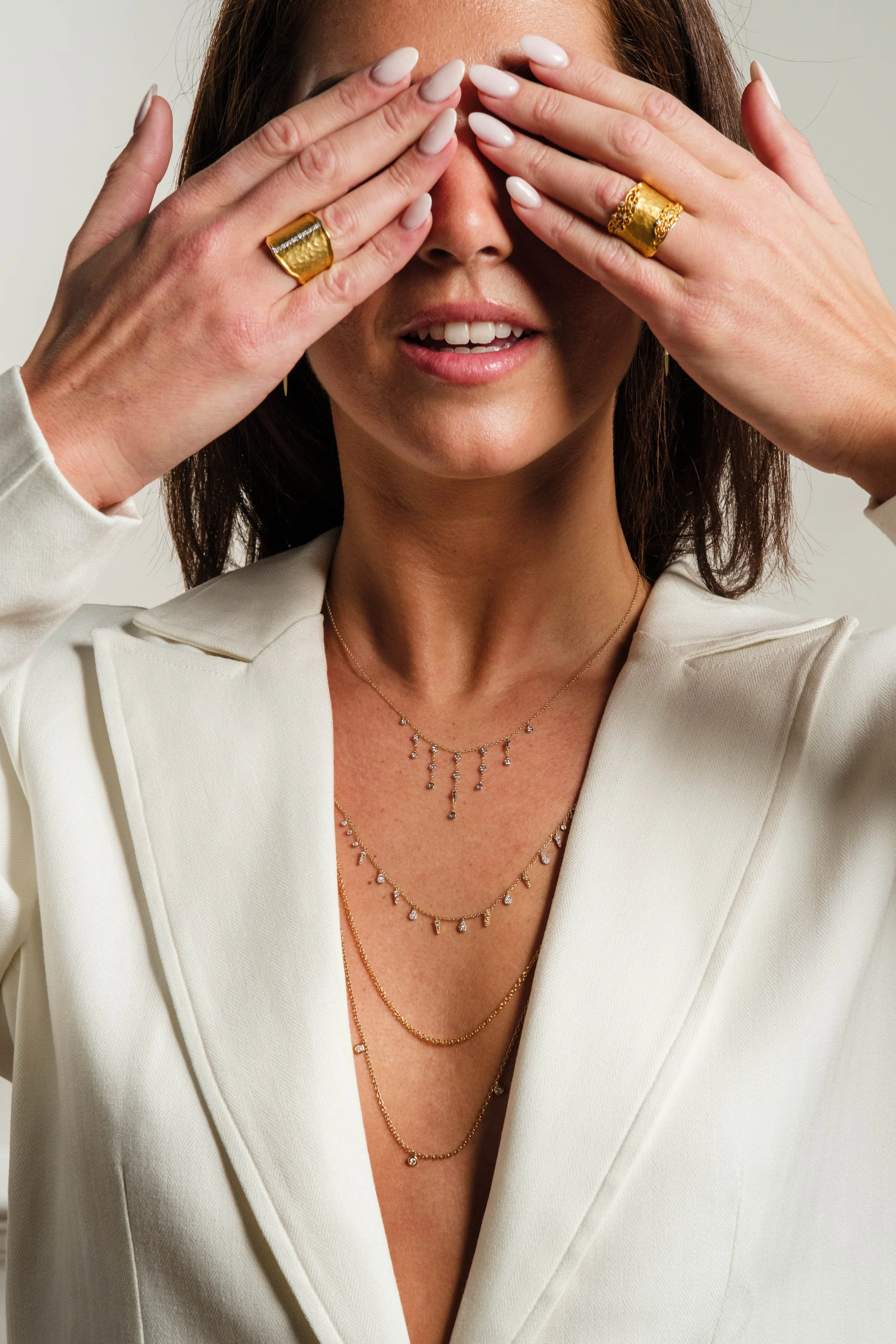 A woman with reddish-brown hair, wearing a white blazer, several gold necklaces with pendant charms, and large gold rings, covering her eyes with her hands which have painted white nails. She is smiling slightly, showing her teeth.