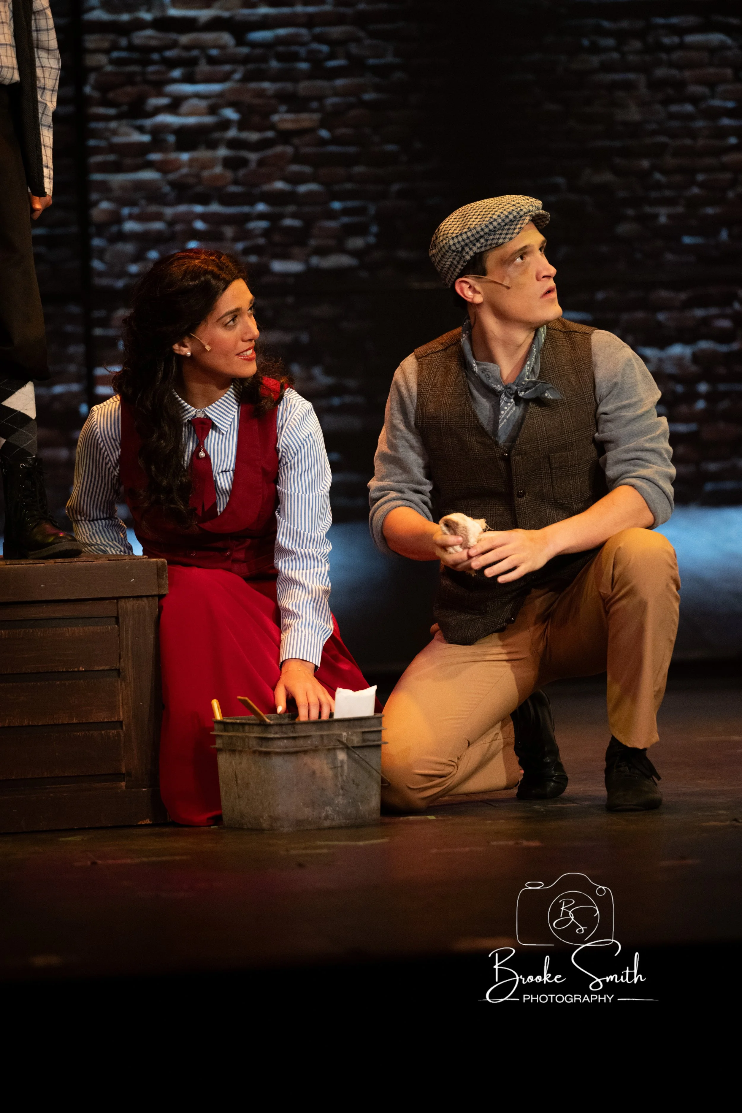 Two actors on stage, a woman with dark hair in a red dress with a striped shirt and a man in a flat cap with a scarf and vest, sitting near a metal bucket, with a brick wall background, appearing to be in a theatrical performance.