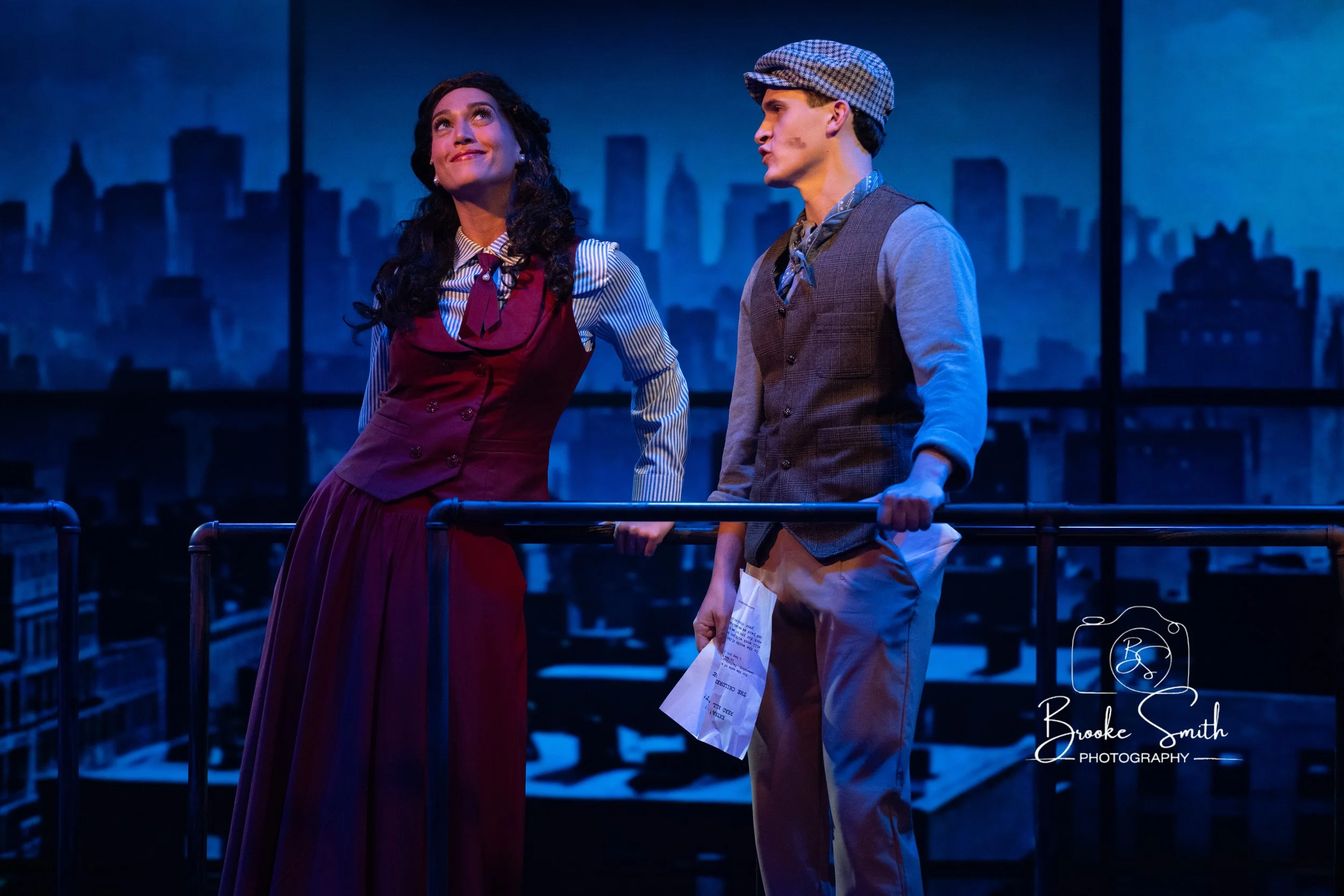 Two actors on stage, a woman and a man, performing in a theater. The woman is smiling with her head tilted slightly upward, wearing a striped shirt and a purple waistcoat. The man is looking at the woman with a serious expression, dressed in vintage 