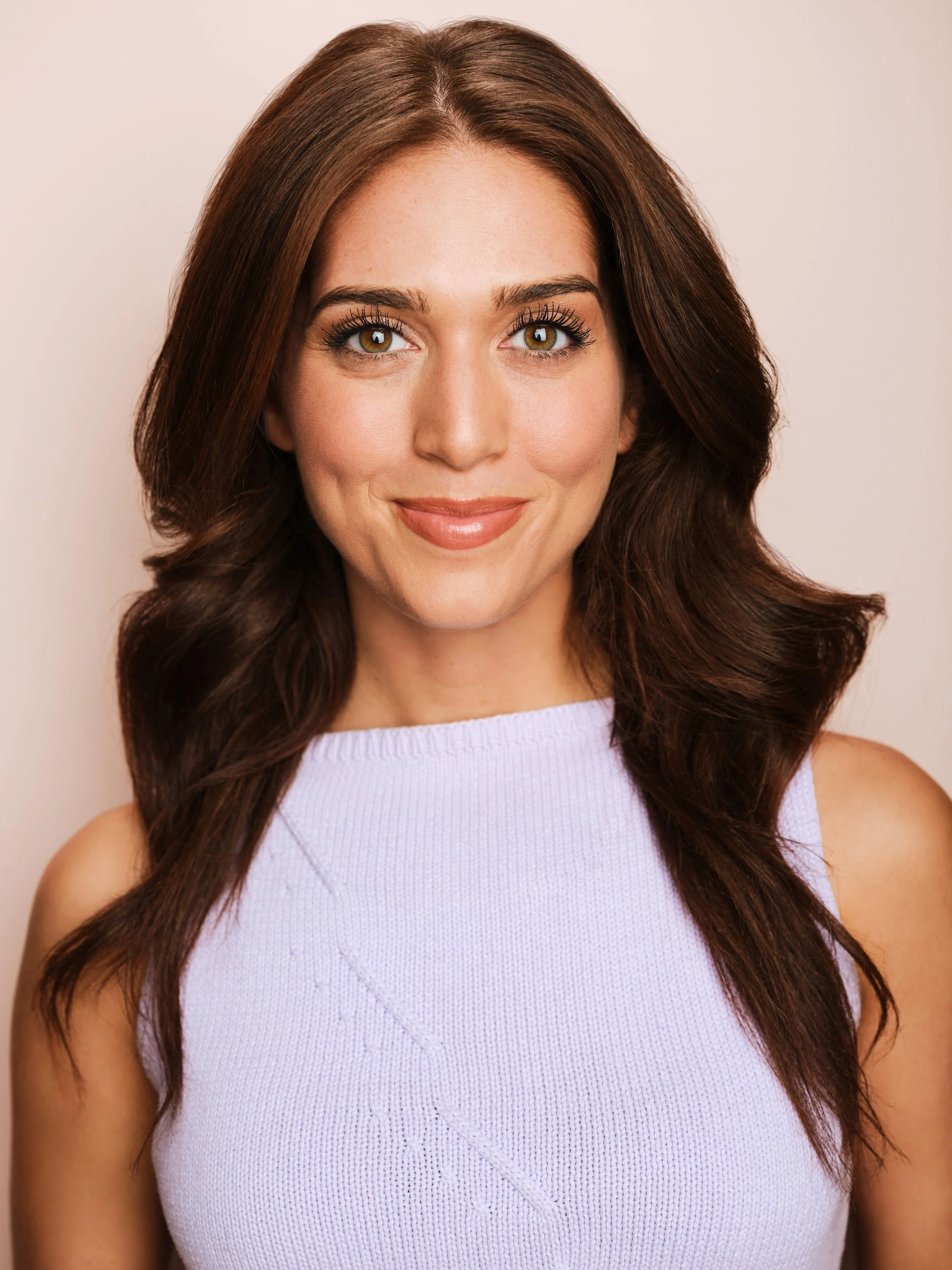 Close-up portrait of a woman with brown hair wearing a sleeveless purple top, smiling softly, against a neutral background.