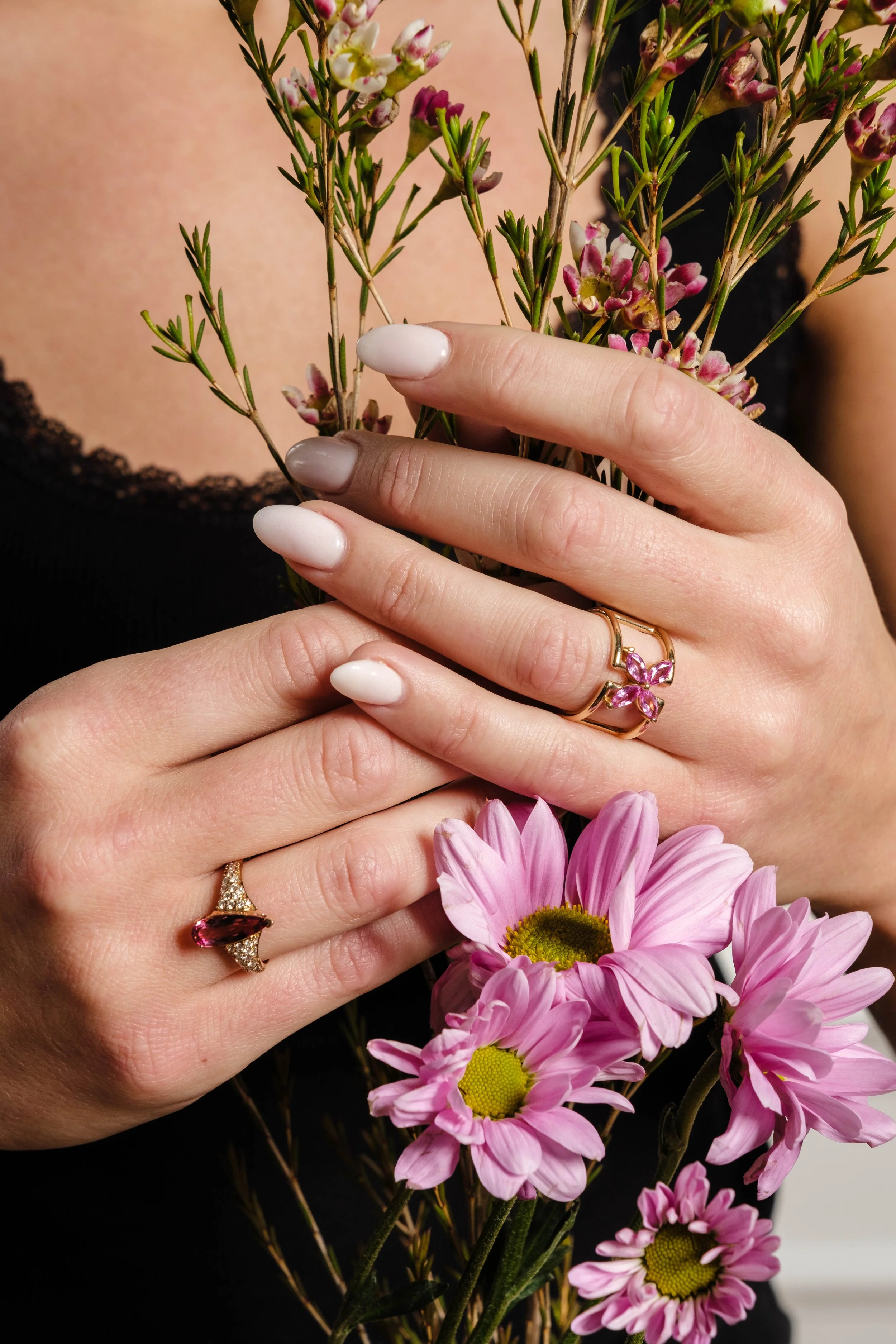 Close-up of hands with manicured nails holding pink flowers and greenery, wearing rings including a pink gemstone ring and a butterfly-shaped ring with pink stones, against a background of pink and white daisies.