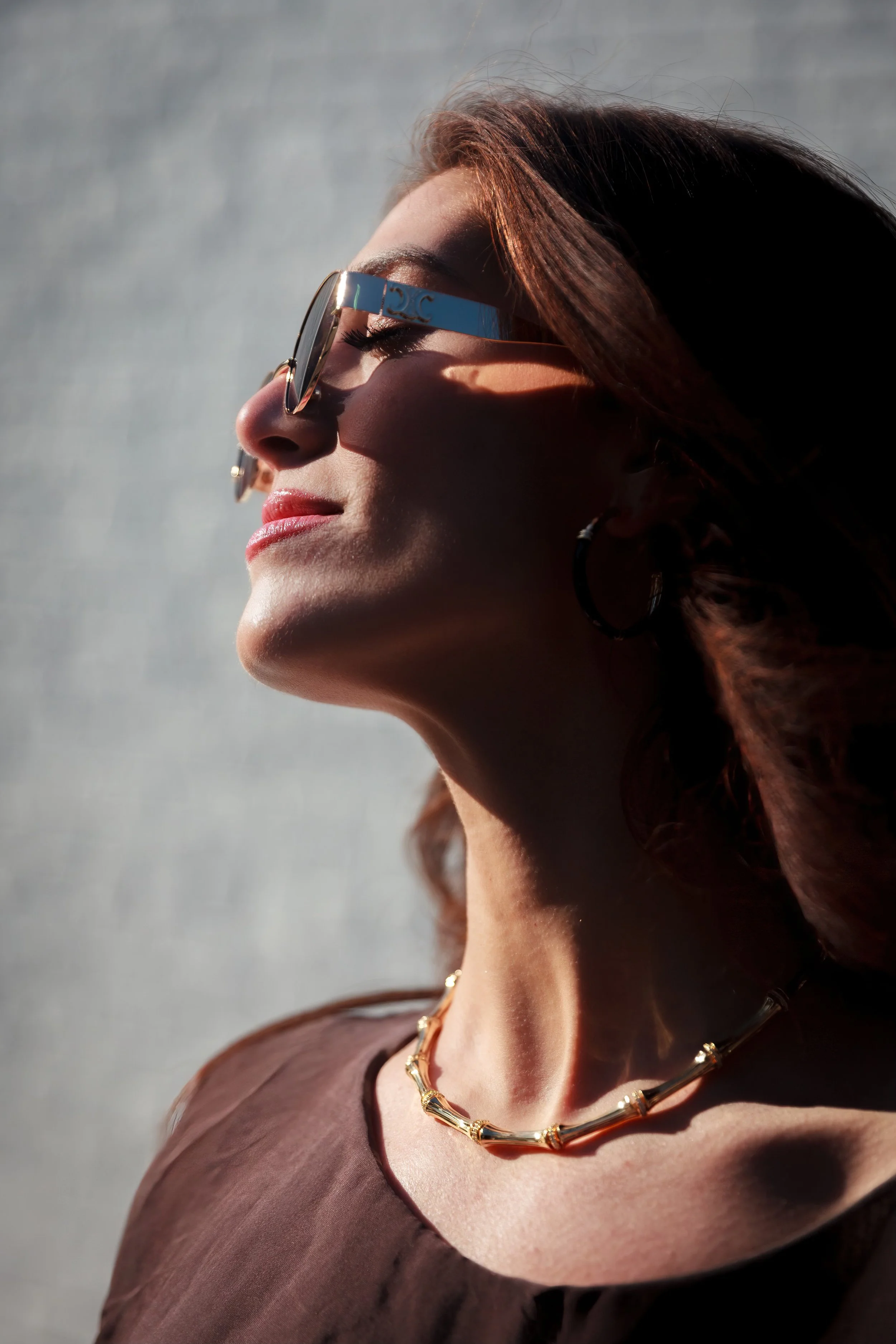 Profile of a woman with red hair wearing large sunglasses, gold jewelry, and a brown top, outdoors in bright sunlight with a gray background.