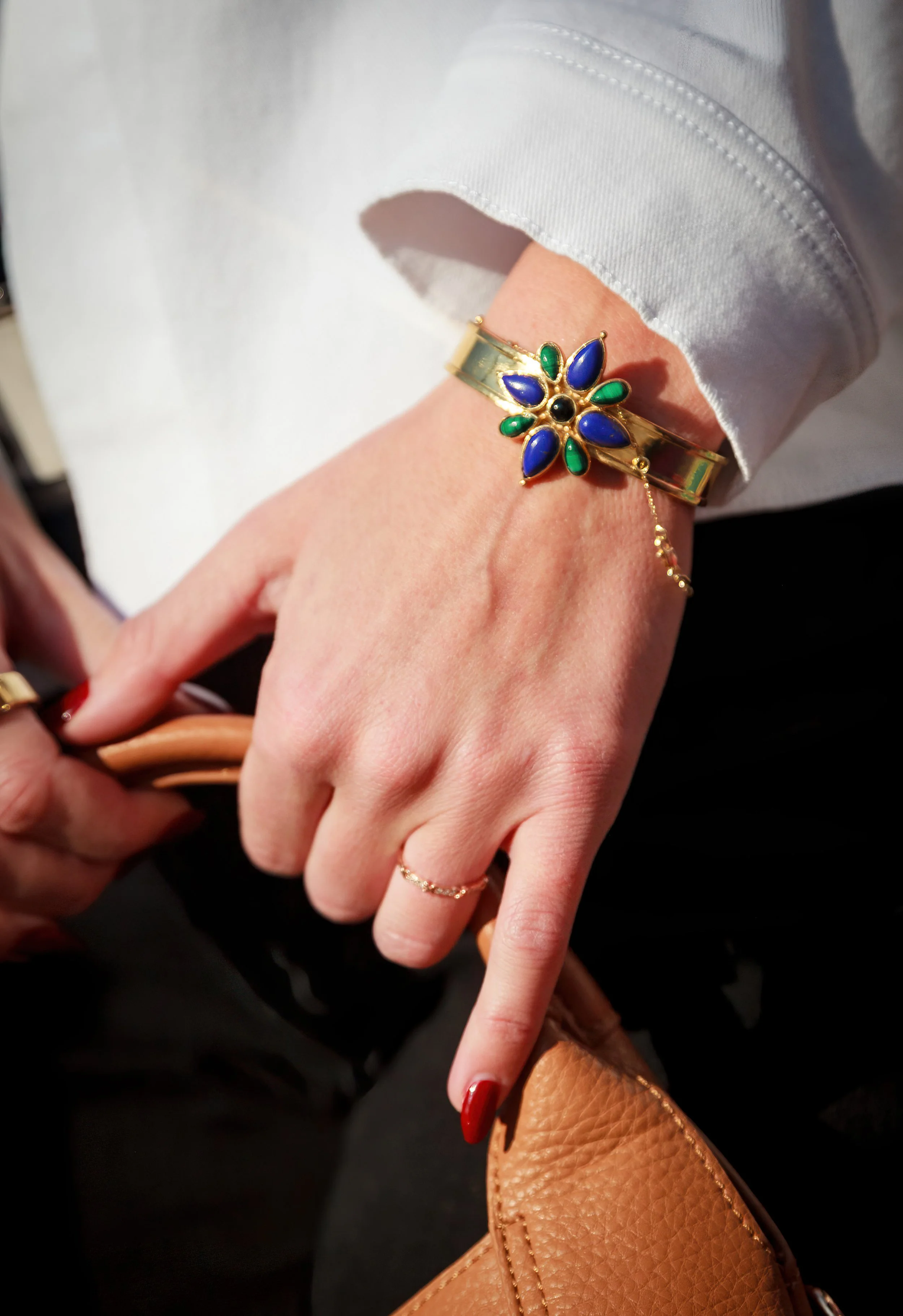 Close-up of a woman's hand and wrist wearing a gold bracelet with a flower-shaped gem design, blue and green stones, and a black center stone, with a brown leather purse in the background.