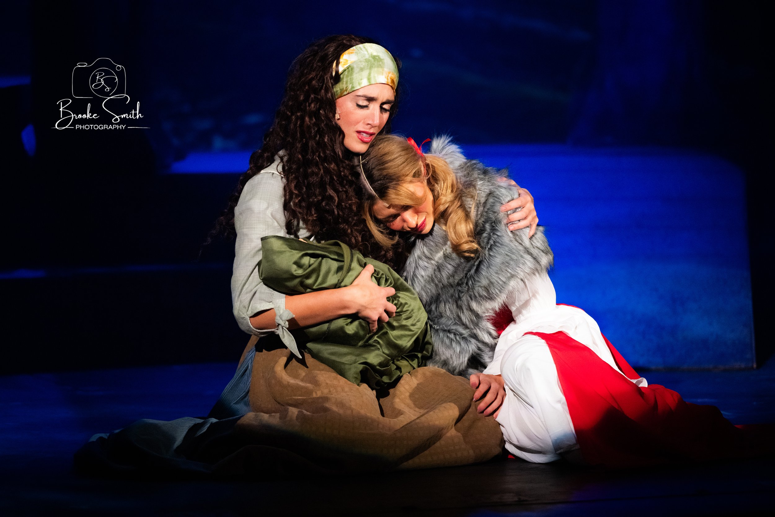 Two women sharing an emotional embrace on stage. One woman has curly dark hair with a green headband, dressed casually, and the other has long blonde hair, wearing a faux fur coat and red and white costume. The scene appears dramatic and heartfelt.