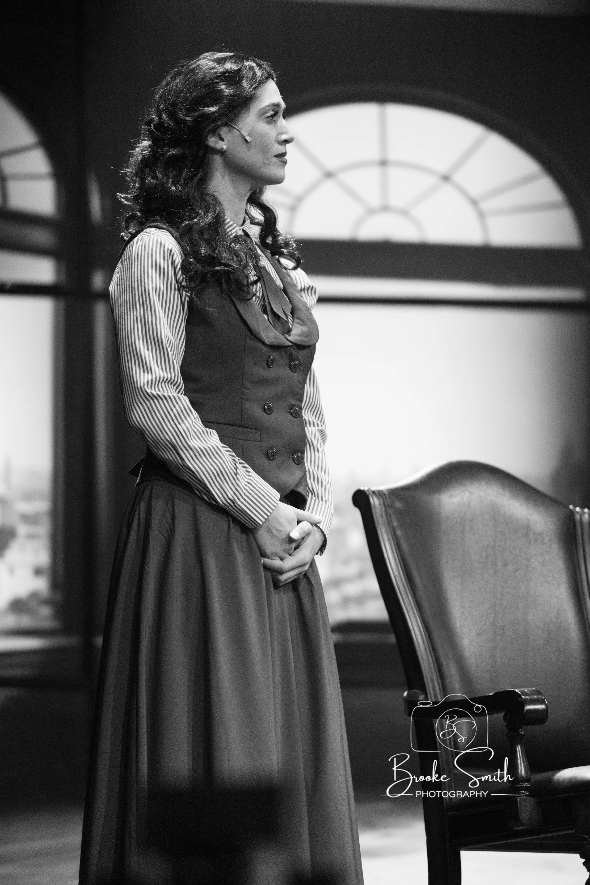 A woman with wavy hair wearing a pinstriped blouse and vest, standing in profile in an indoor setting.