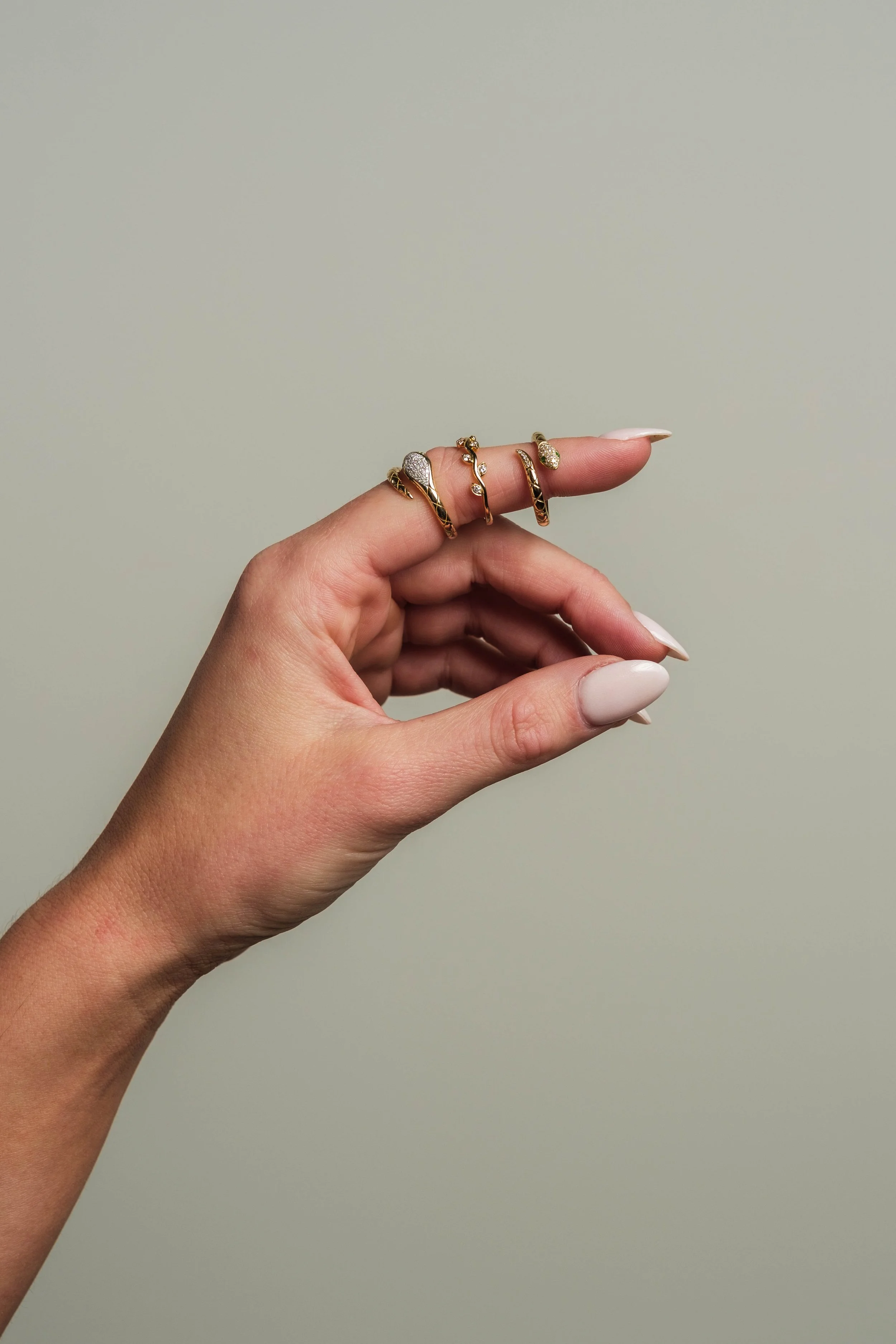 A hand with light skin shows four gold rings on the fingers, decorated with diamonds and intricate designs, set against a plain light beige background.