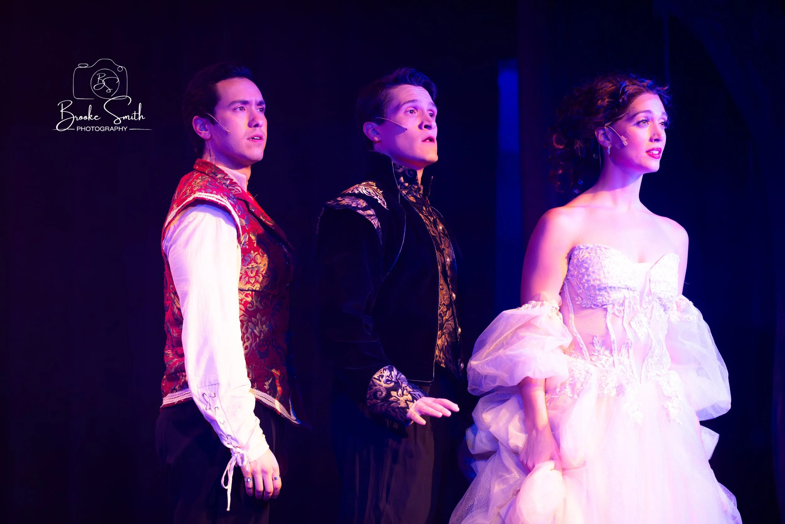 Three actors on stage dressed in period costumes, one man in a red patterned vest and white shirt, another man in black with gold embellishments, and a woman in a white strapless gown with lace details. They are looking in the same direction with exp