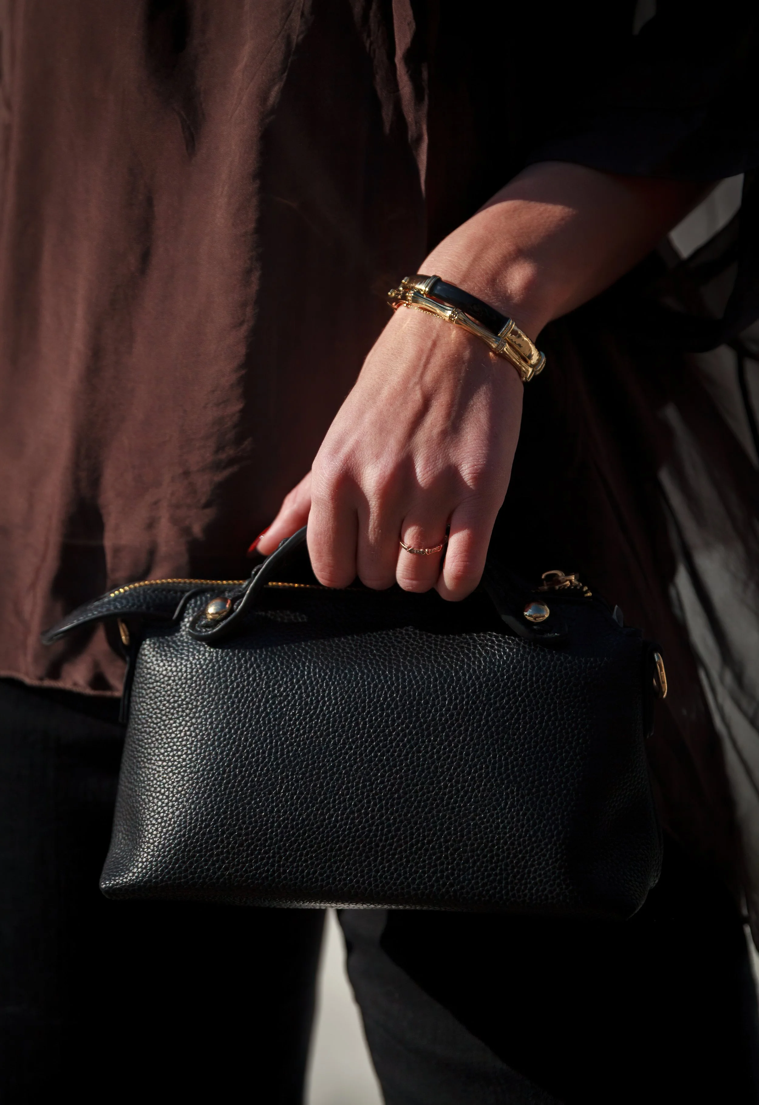 Close-up of a woman's hand holding a black textured purse, wearing gold jewelry, including a bracelet and rings, against dark clothing.