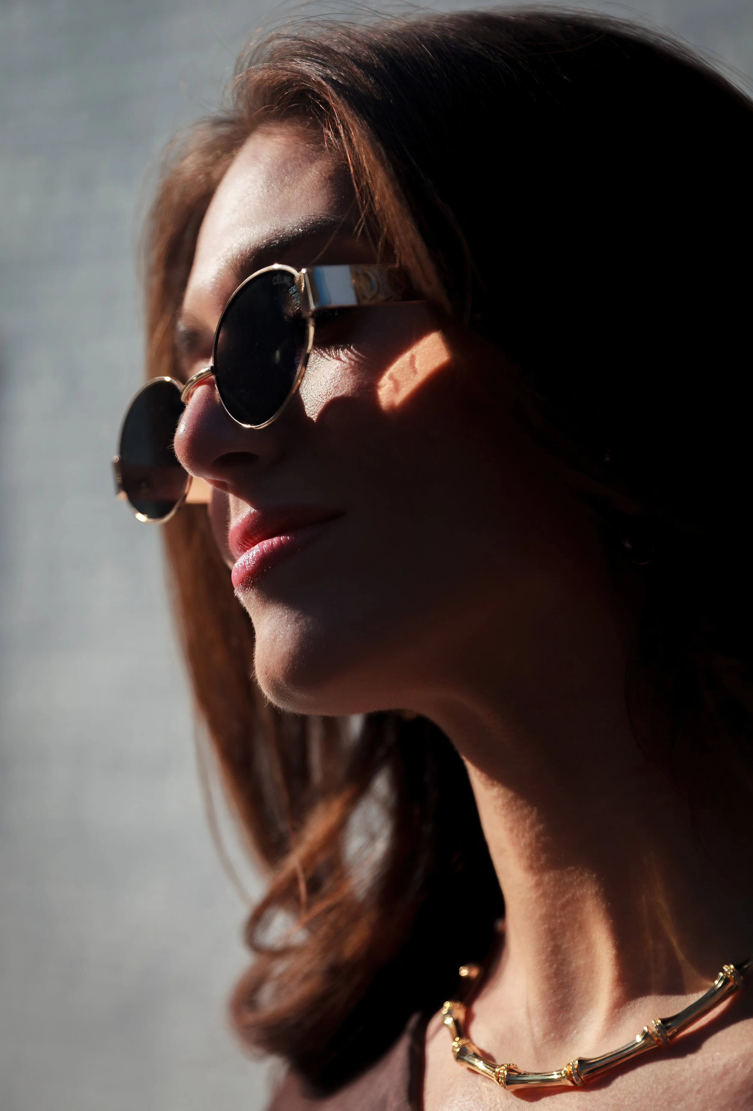 Close-up of a woman with wavy brown hair wearing round sunglasses and a gold chain necklace, with sunlight casting shadows on her face.