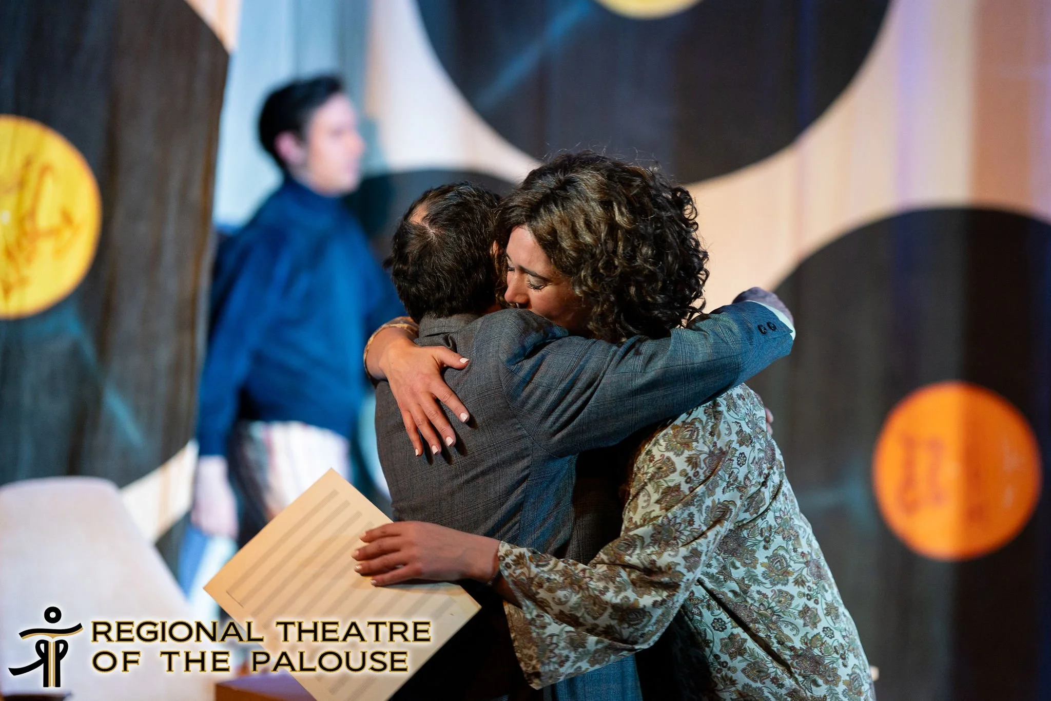 A woman and a young person hug each other in a warm embrace at an event. The woman has curly hair and is wearing a floral blouse, while the young person has short hair and is dressed in a gray suit. Behind them, a blurred background shows a man in a 
